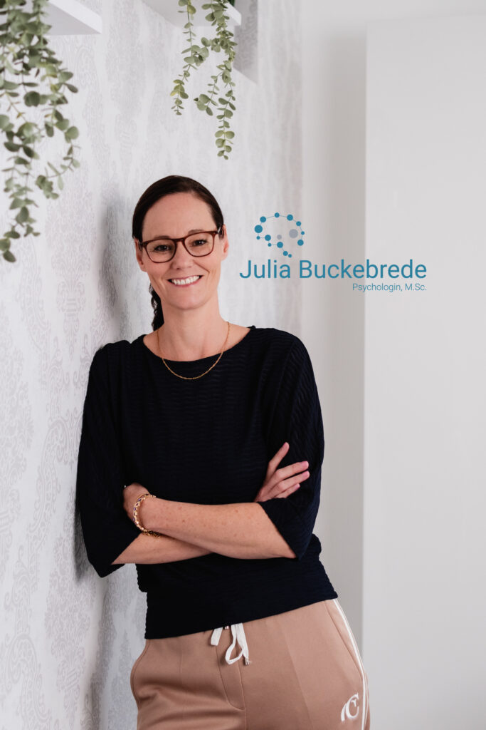 Julia Buckebrede Julia Buckebrede lehnt an einer Wand, hat die Arme verschränkt und lacht